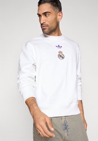 Λευκό φούτερ με στρογγυλή λαιμόκοψη, μοβ λογότυπο Adidas, έμβλημα της Ρεάλ Μαδρίτης με κορώνα, ριμπ μανσέτες και τελείωμα, με άνετη γραμμή.