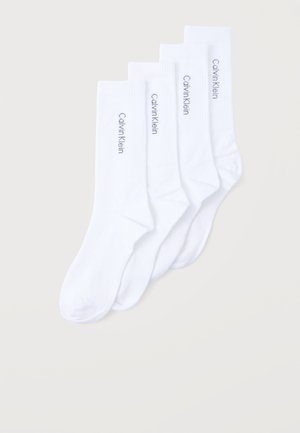 Neljän parin valkoiset nilkkasukat pinottuna, jokaisessa musta "Calvin Klein" -logo sivukahvassa yksinkertaisen vaalean taustan edessä.