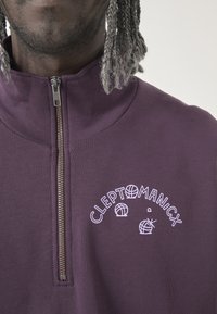 Lila Zip-Hoodie mit weißer Aufschrift "CLEPTOMANICX" und grafischen Designs, mit samtiger Textur und hohem Kragen.