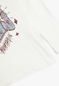 Langærmet hvid bomulds skjorte med blå grafikprint med stjerner, røde detaljer og en sideslids ved sømmen.
