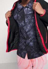 Fleecová bunda s červeným lemem, otevřená, aby odhalila košili s černým a purpurovým vzorem pod ní. Košile má límec a zapínání na knoflíky.