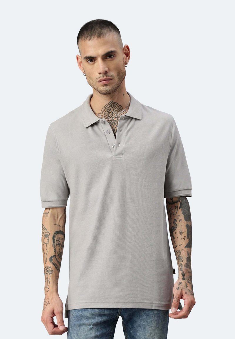 Polo grigio a maniche corte, con patta a due bottoni e tessuto testurizzato. Indossato con jeans blu, che mostrano tatuaggi dettagliati sulle braccia.