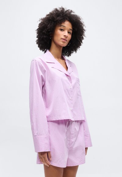 Γυναίκα με κατσαρά μαλλιά, που φοράει ροζ και λευκόριγέ πουκάμισο πιτζάμας με κουμπιά και αντίστοιχα σορτς, στέκεται μπροστά σε απλό φόντο.