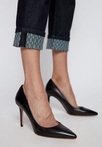 Zapatos de tacón alto negros de piel con punta afilada y tacón de aguja, combinados con pantalones de mezclilla oscuros con dobladillos, que tienen un forro de patrón azul.