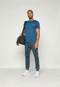 Blå treningsskjorte med teksturert finish, kombinert med mørkegrønne joggebukser med hvite sidestreker. En svart treningsbag holdes ved siden.