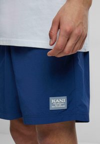 Tamsiai mėlynos šortai iš lengvos medžiagos su stačiakampiu logotipo pažymėjimu "KANI" apatiniame kairiajame kampe. Paprastas dizainas ir švarios linijos.