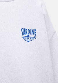 Harmaa puuvillaneule, jossa on sininen painettu teksti "SARDINE" ja kalapiirros vasemmassa rinnassa. Sujuva tekstuuri, rento istuvuus.