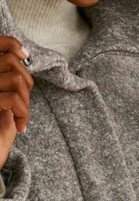 Manteau en laine texturé gris avec une fermeture à bouton, présentant un col doux. Une main soulève délicatement le col, révélant un pull côtelé beige clair en dessous.