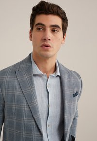 Helles blaues Karo-Blazer aus strukturiertem Stoff mit spitzen Revers, getragen über einem hellblauen Hemd mit Knopfleiste. Enthält ein Einstecktuch.