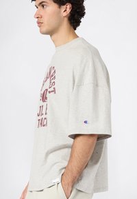 Zijaanzicht van een man in een losse lichtgrijze T-shirt met bordeauxrode tekst en logo's, met de handen in de zakken, op een effen witte achtergrond.