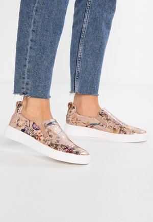 Ben iført blå korte jeans og slip-on sneakers med skinnende beige blomsterprint og hvide tykke såler.