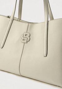 Beżowa torba typu tote ze skóry o teksturowanej powierzchni, z podwójnymi uchwytami i wyraźnym emblematem logo z przodu. Prosty design, bez widocznych metalowych elementów.