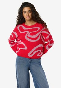 Rdeč pulover z roza abstraktnimi vzorci zavojčkov, pleten teksturo, okrogel izrez in dolgimi rokavi, kombiniran s svetlo modrimi širokimi kavbojkami.