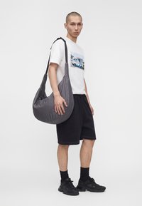 Jeune homme portant un t-shirt blanc avec un motif graphique, un short noir, des baskets noires, et portant un grand sac à bandoulière gris sur un fond uni.