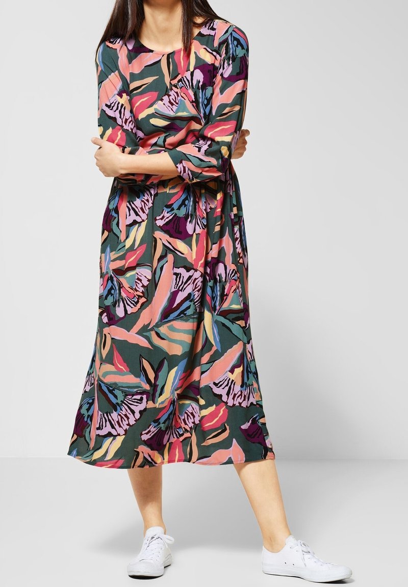 Midi šaty s květinovým vzorem a tříčtvrtečními rukávy; obsahují zelenou, růžovou, fialovou a žlutou barvu. Nosené s bílými teniskami.
