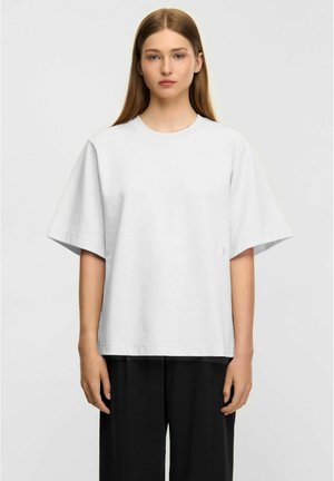 Ung person med lyst, glat, lyst brunt hår iført en oversized, ensfarvet hvid kortærmet t-shirt og løstsiddende sorte bukser mod hvid baggrund.