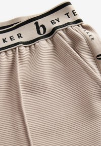 Beige ribbet stoff med en blød tekstur, prydet med et sort taljeelastik med "b by TE" branding. Designet er samlet for en tætsiddende pasform.