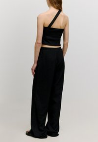 Zwarte crop top met één schouderband, gecombineerd met wijde zwarte broek. De stof lijkt gestructureerd, met een ontspannen pasvorm in de gehele outfit.