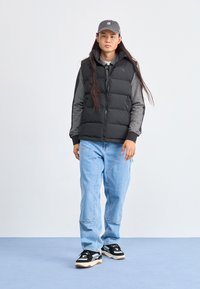 Gilet rembourré noir, chemise à manches longues grise, jeans bleu ample et baskets noires avec des accents blancs. Le modèle porte une casquette grise. Fond neutre.