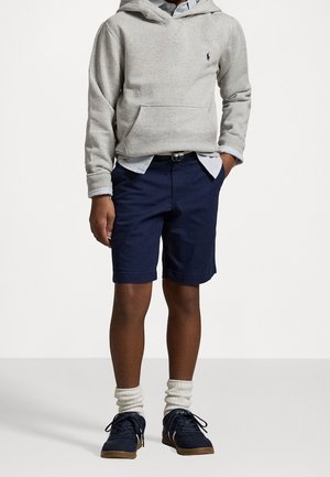 Sudadera gris con capucha y bolsillo delantero, pantalones cortos azul marino y zapatillas azul marino. Calcetines blancos hasta el tobillo aportan textura. Se ve el dobladillo de una camiseta a rayas.