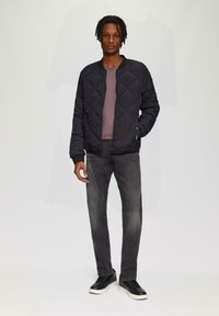 Zwarte gewatteerde bomberjack met een ronde hals, gecombineerd met een mauve shirt en donkere jeans; zwarte sneakers met witte zolen.