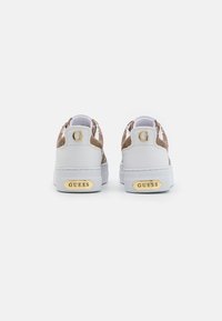 Witte sneakers met een gestructureerde goudbruine achterkant, voorzien van een opvallende gouden "G" en "GUESS" logo op de hak, dikke witte rubberen zool.
