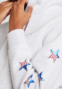Mani che sistemano il colletto di una felpa leggera grigia decorata con stelle che presenta il motivo della bandiera americana sulle maniche.