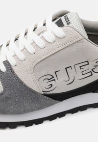 Baskets grises et beiges avec des matériaux en daim et en mesh, lacets blancs, accents texturés et un logo noir sur le côté. Semelle extérieure en caoutchouc.