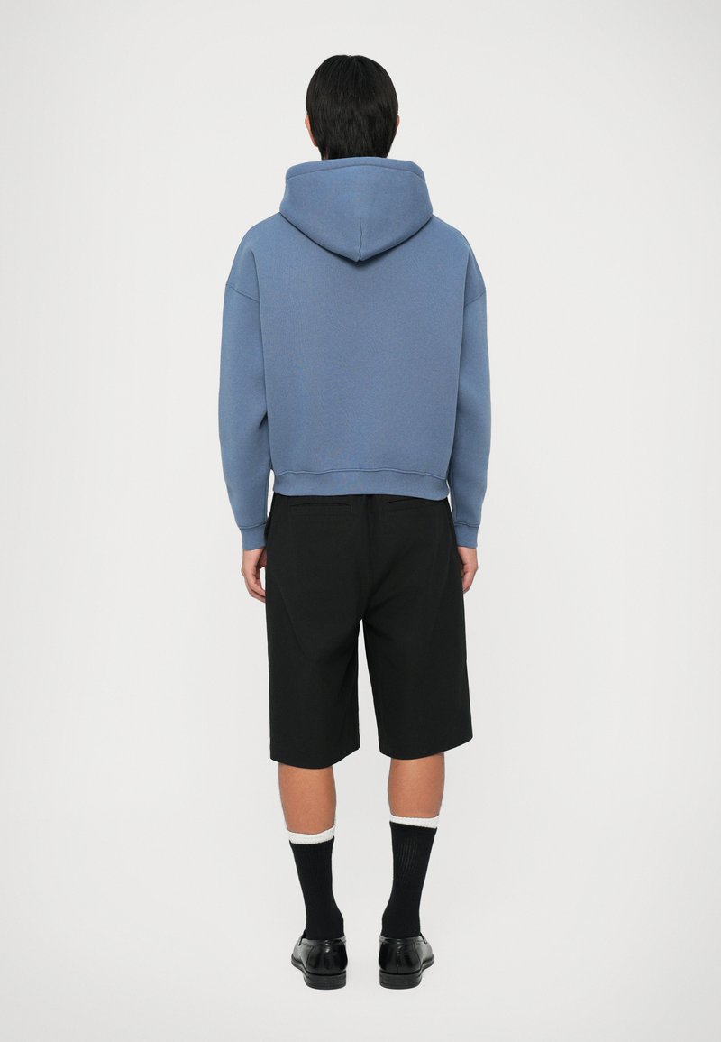 Persona in piedi di spalle, che indossa un felpa blu, pantaloni corti neri fino al ginocchio, calze nere con rifiniture bianche e scarpe eleganti nere.