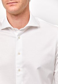 Weißes Hemd mit Knopfleiste, klassischem Kragen, weichem Textil und Knöpfen aus Perlmutt. Das Design ist figurnah und der Stoff ist glatt.