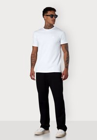 T-shirt blanche à manches courtes avec un col ras-du-cou et un petit logo sur la poitrine, associé à un pantalon noir et des sneakers blanches.