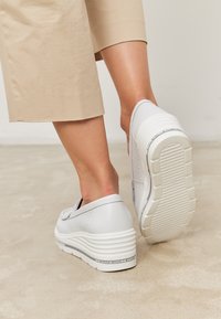 Sneaker bianca con tomaia in materiale sintetico liscio, design slip-on, perforazioni testurizzate e una suola spessa e scanalata con un accento argentato.