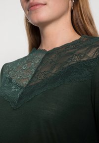 Tmavě zelená košile se krajkovou vrstvou na výstřihu. Krajka má květinový vzor a vlnité okraje, což přidává texturu a detail.