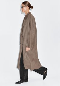 Lang bruine wollen jas met een lulkraag, twee knopen en een oversized pasvorm. Gecombineerd met zwarte jeans en puntige zwarte leren schoenen.