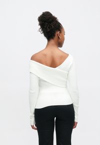 Haut blanc à épaules dénudées en maille côtelée avec manches longues, présentant un dos drapé. Associé à un pantalon noir ajusté.