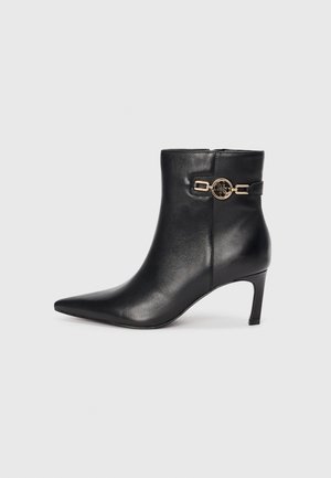 Bottine en cuir noir avec un bout pointu, un talon haut et fin, et un accent doré avec un design circulaire et un détail de sangle.
