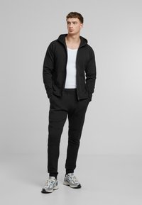 Svart zip-up huvtröja med dubbel-lager huva, i kombination med svarta tapered joggers. Bärs över en vit tanktop och silverfärgade sneakers.