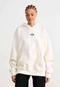 Hoodie sovrastante di colore crema realizzato in tessuto morbido, con tasca frontale e un piccolo logo nero sul petto. Maniche lunghe con estremità a costine.