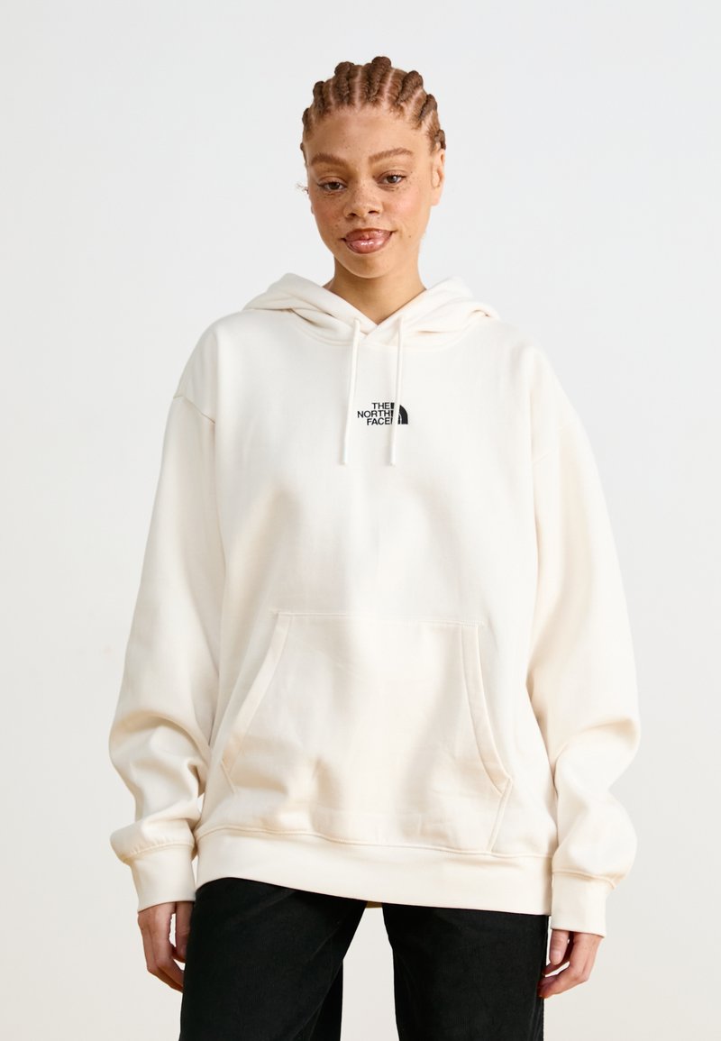 Hoodie sovrastante di colore crema realizzato in tessuto morbido, con tasca frontale e un piccolo logo nero sul petto. Maniche lunghe con estremità a costine.