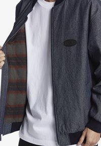 Denim bomberjacka med en mörkblå utsida, svart logotyp patch och en rutigt foder i röda och gröna nyanser. Har dragkedja och ribbade ärmslut.