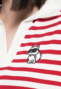Weißes Poloshirt mit roten horizontalen Streifen, mit einem gestickten cartoonhaften Kater, der eine Sonnenbrille trägt, auf der linken Seite.