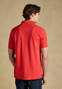 Homme aux cheveux foncés portant un polo rouge à manches courtes et un jean sombre, vu de dos sur un fond neutre.