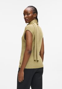 Strikket ærmeløs top i guld med en høj krave og draperet tørklædedetalje, parret med mørke bukser. Struktureret stof og synlige knapdetaljer på siden.