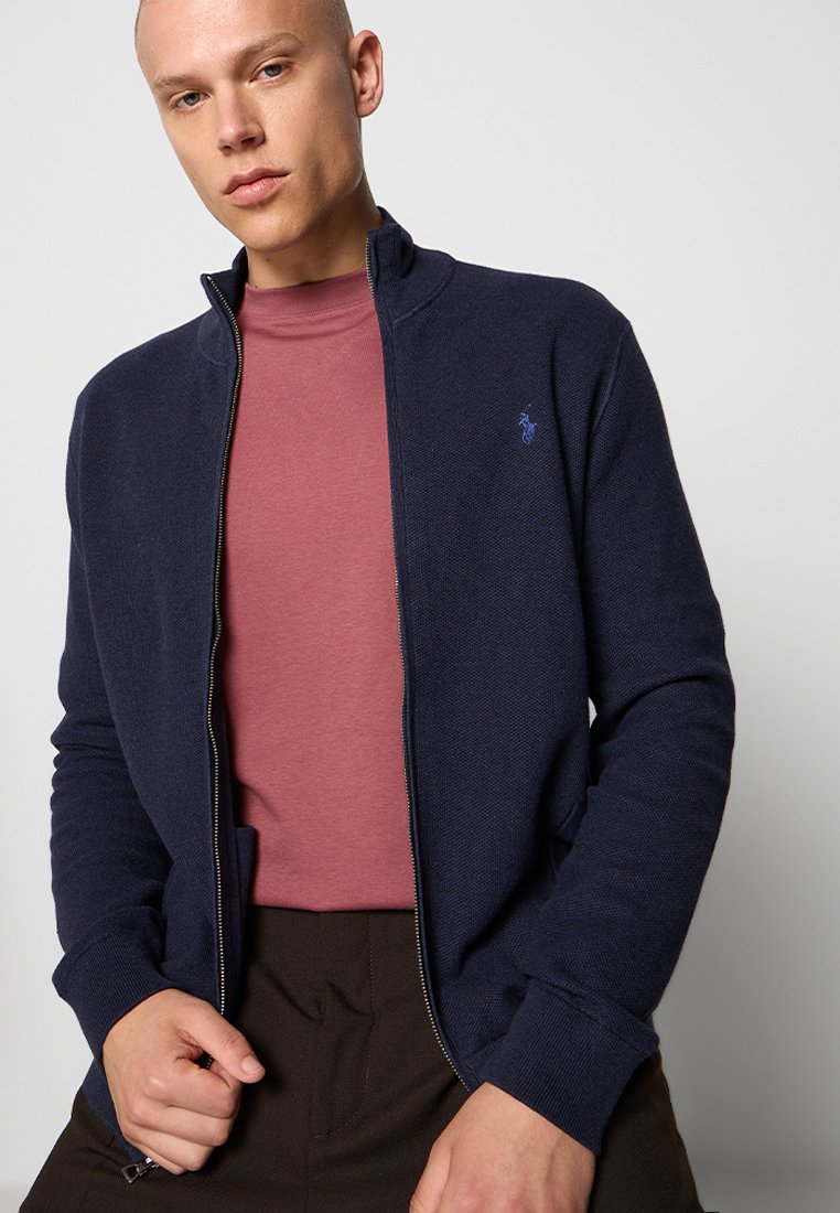 Marineblaue Zip-Jacke mit strukturiertem Strick, hohem Kragen und dezentem Logoakzent. Getragen über einem burgunderfarbenen Rundhals-Shirt.