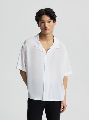 Giovane uomo con capelli scuri che indossa una camicia bianca ampia a maniche corte con bottoni e pantaloni neri, in piedi davanti a uno sfondo grigio chiaro.