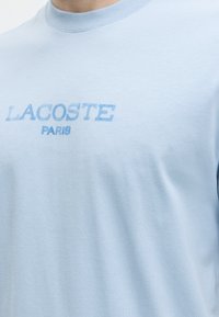 T-shirt bleu clair col rond avec l'inscription "LACOSTE PARIS" imprimée en bleu délavé sur la poitrine.
