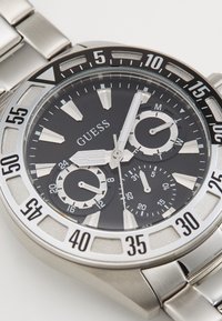 Stříbrné kovové náramkové hodinky s černým ciferníkem, třemi subčíselníky pro 24 hodin, den a datum a velkými bílými ručičkami ukazujícími čas 9:05:02.