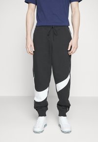 Pantalones de chándal negros con acentos de panel curvado en blanco, cintura elástica y puños. El material parece suave; combinados con una camiseta de color marino y zapatillas blancas.