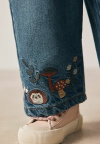Denim-Hosenbein mit bestickten Pilzen, Blättern, Blumen und einem Igel über einem beigen Sneaker mit gemusterten Schnürsenkeln.
