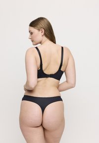 Kobieta z długimi brązowymi włosami, ubrana w czarny stanik i stringi, pokazana od tyłu na jednolitym jasnym tle.